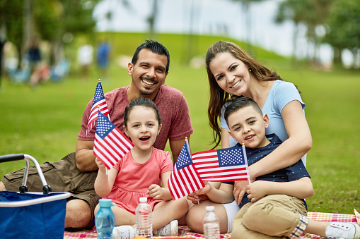 Smiling Hispanic American parents in late 30s with 4 and 6 year old children holding American flags and celebrating holiday with picnic at Miami park.