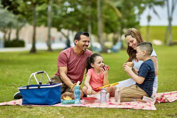 Hispanic American parents in late 30s enjoying picnic lunch and family time with their young children at Miami public park.
