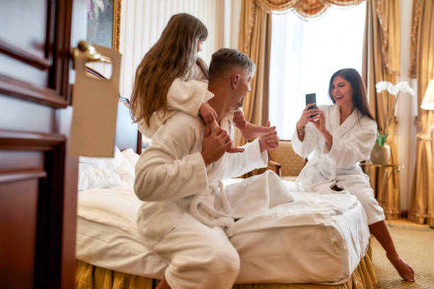 sorriso. genitori e figlia in accappatoi bianchi che trascorrono la mattina insieme. papà e bambini sono in posa per una foto seduti sul letto nella lussuosa camera d'albergo. famiglia, viaggi, resort, concetto di vacanza - hotel wireless technology bedroom hotel room foto e immagini stock