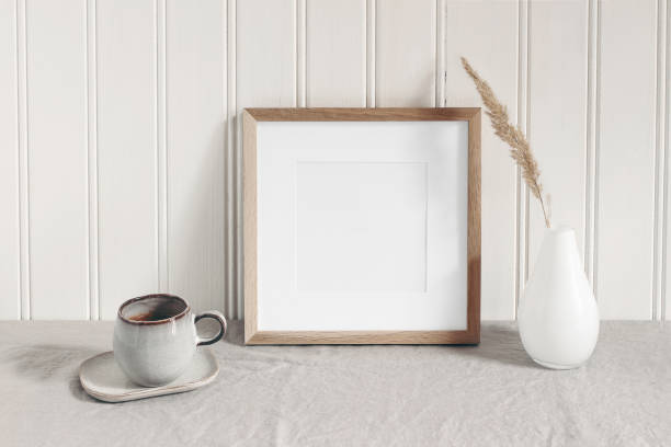 istock Square empty wooden frame mockup with modern ceramic vase, dry grass, cup of coffee on table. White beadboard wainscot wall paneling background. Scandinavian interior, home design. Art concept. 1209745944