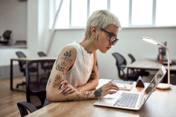 concentré au travail. vue de côté de jeune et attrayante femme d’affaires blonde tatouée dans des lunettes utilisant l’ordinateur portatif tout en travaillant dans le bureau moderne - tatouage femme photos et images de collection