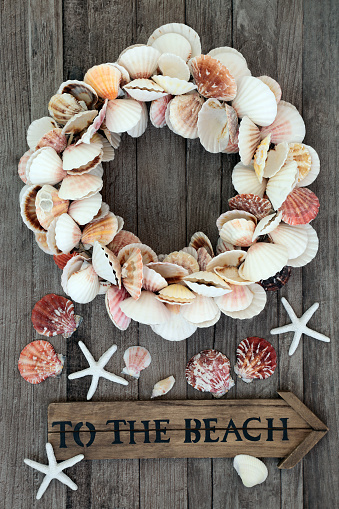 Heart shaped wreath made of scallop seashells on rustic wood background with old to the beach sign & loose shells. Romantic symbol and summer holiday theme concept.