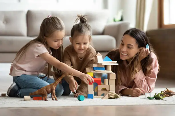 Loving young mom lying on warm home floor with cute small daughters construct with building wooden bricks, happy mum have fun playing with little girl kids in living room, relaxing on weekend together