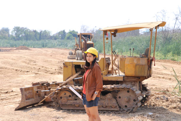 istock Female civil engineer or architect with yellow helmet standing and turn sideways with Bulldozer Truck with Front Loader Truck Engineering Construction Car Vehicle. 1209789439
