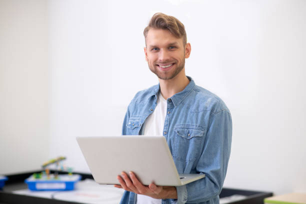 istock Male teacher standing, holding laptop and smiling 1209797819