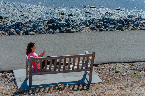 Woman sitting on a bench in a park close to a lake checking her phone