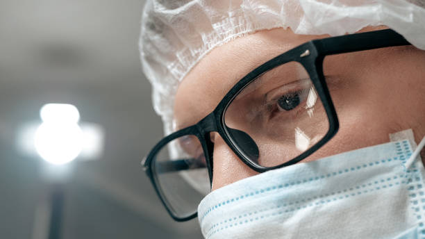 istock Man surgeon with glasses in surgical mask looks down at patient. Operation, practitioner, operating room, surgery, transplantation, medicine concept. Close-up 1209817290
