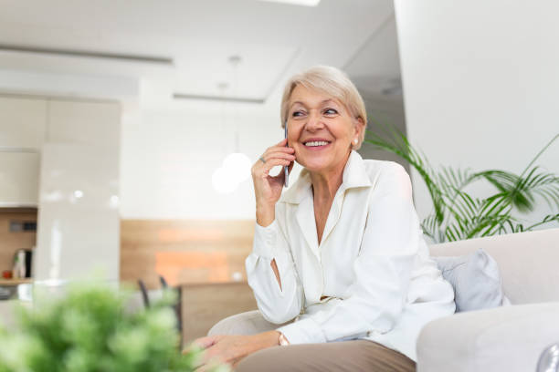 belle femme âgée caucasienne ayant la conversation de téléphone avec son vieil ami utilisant l’appareil électronique, discutant des dernières nouvelles avec le sourire mignon sur son visage, s’asseyant à la maison - senior adult independence lifestyles home interior photos et images de collection