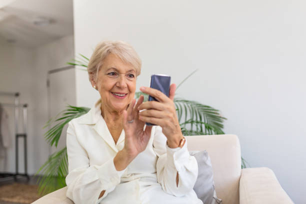 donna matura con il cellulare sulle mani seduta in camera e che invia messaggi ai suoi amici e familiari - grandparent adult smiling looking at camera foto e immagini stock