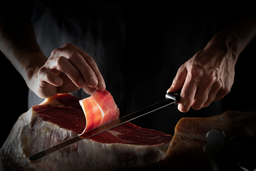 Iberian ham serrano ham slice cutting hands and knife hands on dark background low key