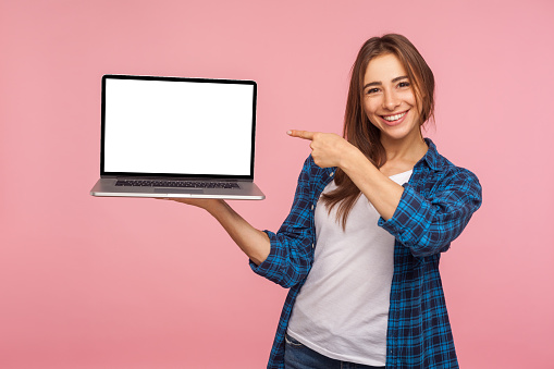 Portrait of happy charming girl in checkered shirt showing blank laptop screen and smiling, pointing mock up, copy space for internet advertising. indoor studio shot isolated on pink background