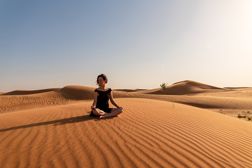 Lady in a black dress in yoga pose on top of the red dune before sunset in windy area without footprints in sand