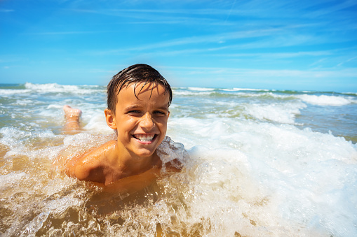 Happy little boy lay on the san of a beach with wave covering him, expressing big cute smile