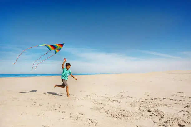Cute boy run on the sand beach near the sea holding colorful rainbow stripped kite
