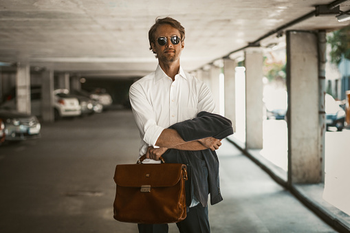 Young sexy businessman standing in a parking lot with his arms crossed over his chest. A young man in a white shirt and blue trousers with sunglasses waiting on his car . Business concept.