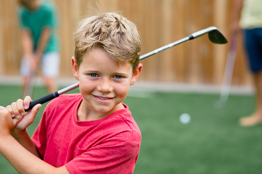 A front view shot of a young boy golfer smiling and looking at the camera, he is holding a golf club.