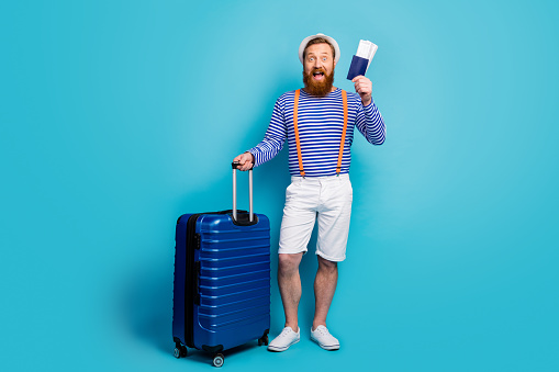 Full body photo of crazy funky man tourist ready go check-in have trolley, bag hold passport scream wear white shorts headwear orange suspenders sun headwear isolated blue color background