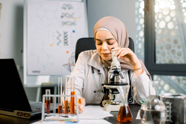 istock Young concentrated female muslim scientist works with microscope and test tubes during experiment in chemical laboratory, sitting at the table and making notes about results of research 1209871753