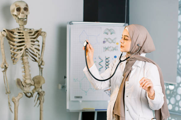 belle jeune femme musulmane du moyen-orient étudiant ou docteur posant à l’hôpital ou à la salle de classe avec le stéthoscope. squelette humain et planche de classe sur le fond - anatomy classroom human skeleton student photos et images de collection