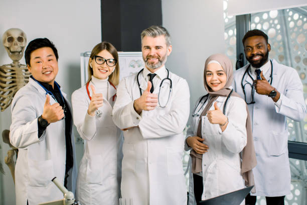 retrato de médico maduro masculino con equipo médico multiétnico, sonriendo y mostrando los pulgares hacia arriba, contra el laboratorio moderno y el esqueleto humano de fondo. concepto de trabajo en ciencia, salud y equipo - scientist lab coat doctor male fotografías e imágenes de stock