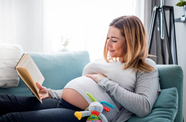 istock Pregnant woman relaxing on sofa reading book looking at belly 1209874210