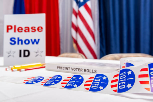 A roll of “I VOTED” stickers on a empty US election registration desk with PLEASE SHOW ID sign A roll of “I VOTED” stickers sitting on the table of an empty registration desk at a US polling station on election day with a “PLEASE SHOW ID” sign, red, white and blue bunting an American flag in the background. voter registration photos stock pictures, royalty-free photos & images