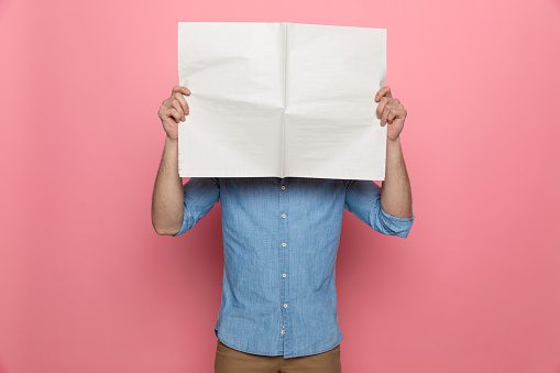 young casual guy covering face with newspaper and standing on pink background