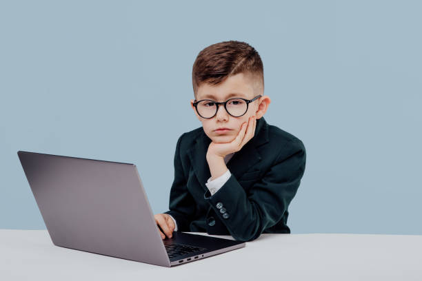 istock exhausted schoolboy with glasses using a laptop, little boss looks at camera isolated on blue background, in studio 1209885733