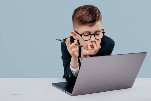 little boy in glasses and suit reading data from laptop sitting at table and working against blue background