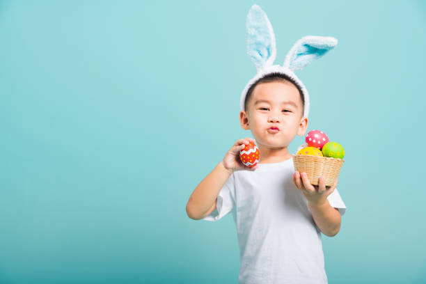 il simpatico bambino asiatico sorride raggiante indossando orecchie da coniglietto e una maglietta bianca, in piedi per tenere un cesto con uova di pasqua complete. e l'altra mano tiene un uovo di pasqua - costume da coniglietto foto e immagini stock