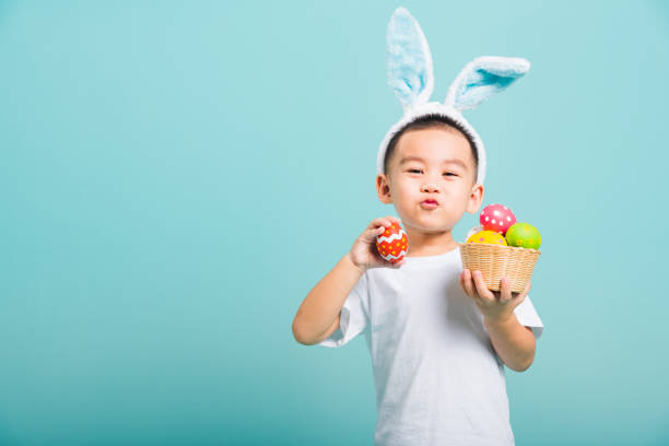 azjatycki cute little boy dziecko uśmiech rozpromieniony sobie uszy króliczka i biały t-shirt, stojący trzymać kosz z pełnymi pisankami. a druga ręka trzyma jajko wielkanocne - easter baby rabbit eggs zdjęcia i obrazy z banku zdjęć