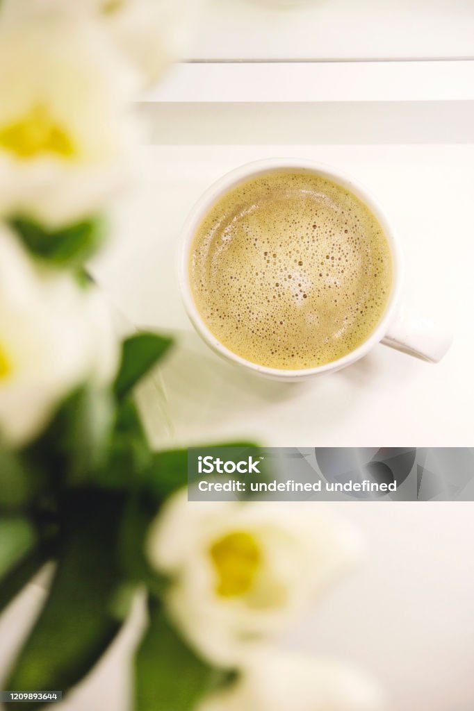 A cup of coffee in the morning next to a bouquet of white tulips. Spring holiday card. White tulips on the table next to a cup of morning coffee in a bright cozy home atmosphere. Congratulations on March 8 and Mother's Day. Above Stock Photo