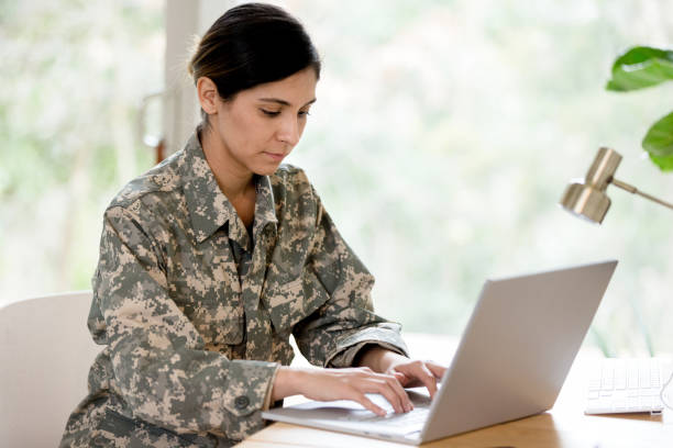 Mid adult female soldier works on laptop at home A mid adult female soldier wearing her uniform works on her laptop at home. camouflage clothing photos stock pictures, royalty-free photos & images