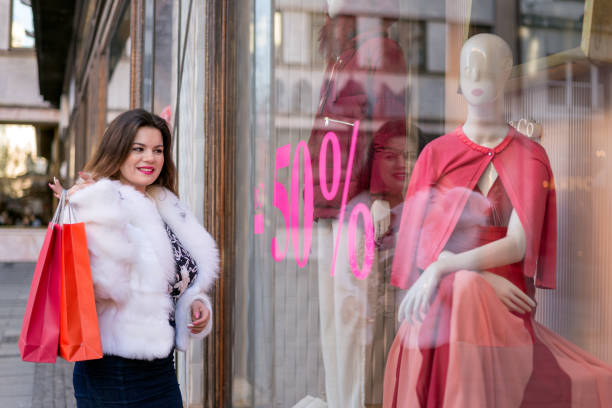 Happy fancy dressed young woman holding shopping bags and looking at the window shop. Shopping, sale, lifestyle concept. stock photo