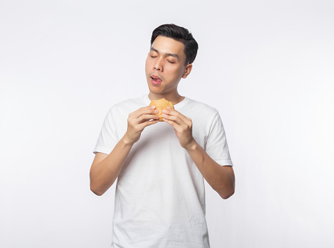 Young Asian man in white t-shirt eating hamburger with happy face isolated over white background.