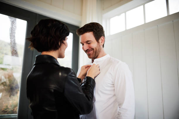 Young woman helping her smiling husband button his shirt Young woman helping her smiling husband with his shirt buttons while getting dressed up at home buttoning stock pictures, royalty-free photos & images