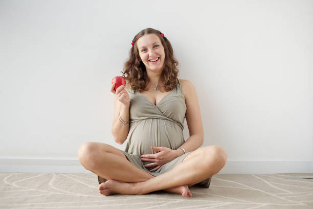 Portrait of happy pregnant woman sitting on the carpet, touching her big belly, holding an apple and laughing. Image of laughing pregnant woman sitting on the floor and holding an apple. Cheerful pregnant European woman with big tummy is looking to the camera . Pregnancy, health people concept. olivia mum stock pictures, royalty-free photos & images