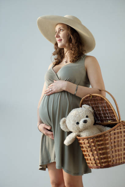 Portrait of the impersonal pregnant woman in the hat holding her big tummy and basket with teddy bear. Motherhood. Image of the smiling pregnant woman touching her big belly. Happy pregnant European woman is waiting for the baby. Pregnancy, motherhood, family, people and expectation concept. White background. olivia mum stock pictures, royalty-free photos & images
