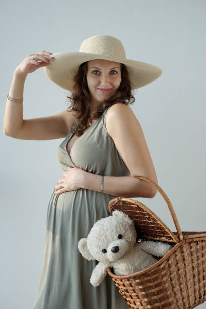 Portrait of the cheerful pregnant woman in the hat holding her big tummy and basket with teddy bear. Motherhood. Image of smiling pregnant woman touching her big belly and basket with teddy bear. Happy pregnant European woman is waiting for the baby. Pregnancy, motherhood, family, people and expectation concept. olivia mum stock pictures, royalty-free photos & images