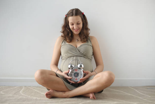 Image of happy smiling pregnant woman, expecting child, sitting on the floor and holding big alarm clock. Happy European woman with big tummy is waiting. Pregnancy, motherhood, people and expectation concept. Waiting for the baby. Motherhood. olivia mum stock pictures, royalty-free photos & images