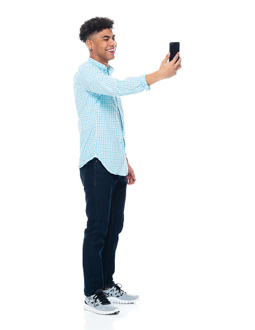 Profile view of aged 18-19 years old with black hair african ethnicity male photography standing in front of white background wearing jeans who is laughing who is photographing and using smart phone