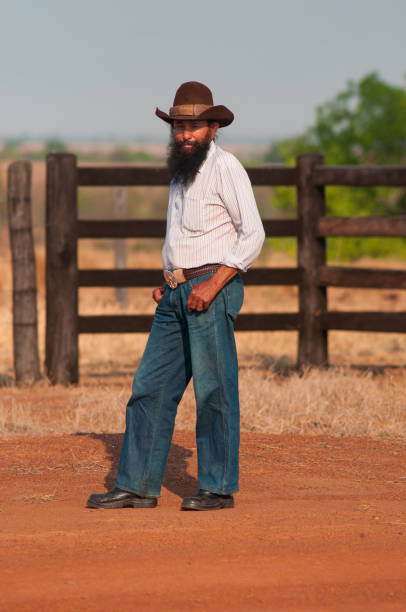 mestiço cowboy em pé na frente do portão de madeira em estrada de terra - 5550 - fotografias e filmes do acervo