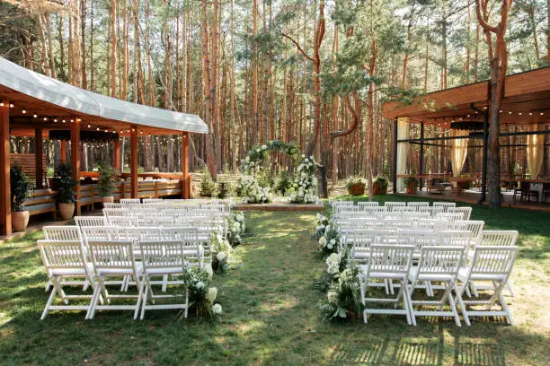 Outdoor Wedding Ceremony. A chic arch in the shape of a ring of white fresh flowers and green branches, white wooden chairs in a row on the grass. Pine forest in the background