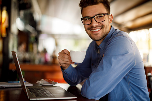 Smiling young man drinking coffee and using laptop at a cafe