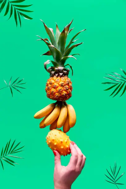 Assortment of tropical fruits, pyramid balancing o human hand on green background. Pineapple, kiwano, kiwi , lichee, banana - tower made of exotic fruits.