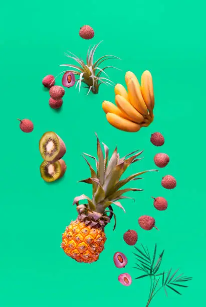 Assortment of tropical fruits and orange smoothie bottle on green background. Pineapple, kiwano, kiwi, lichee and banana - exotic fruits, levitation and vitamin balance.