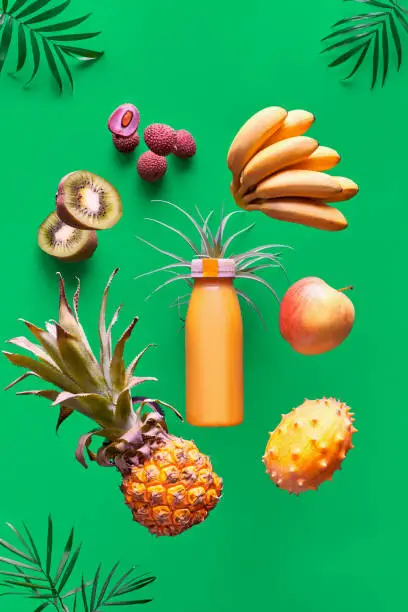 Assortment of tropical fruits and orange smoothie bottle on green background. Pineapple, kiwano, kiwi fruit, lichee and banana - exotic fruits, levitation and balance.