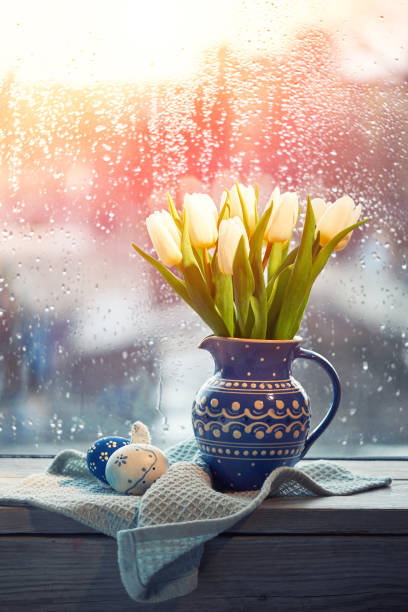 istock Easter decorations in trendy classic blue color. White tulips in traditional ceramic jug, blue Easter eggs with polka dots on a window on rainy day. Toned rustic European Springtime decor, Sun flare 1209966862