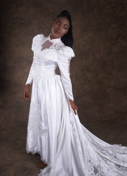 Young Black woman in 1980s style wedding dress, full length with train. Vertical studio shot on brown of young Black woman in 1980s style ornate wedding dress with high collar and long train. Front view holding part of train. high collar stock pictures, royalty-free photos & images