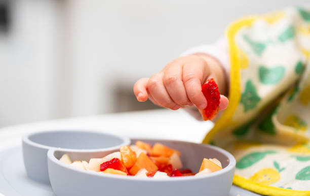 makro nahaufnahme von baby hand mit einem stück früchte sitzen in kinderstuhl kind essen gesunde ernährung - kleinstkind stock-fotos und bilder